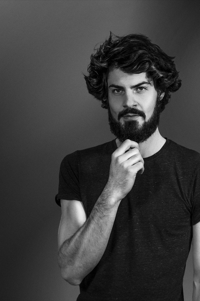 Immanuel Holtmannspoetter, photographed in black and white against a plain grey wall, one hand at his chin, looking directly at the camera.